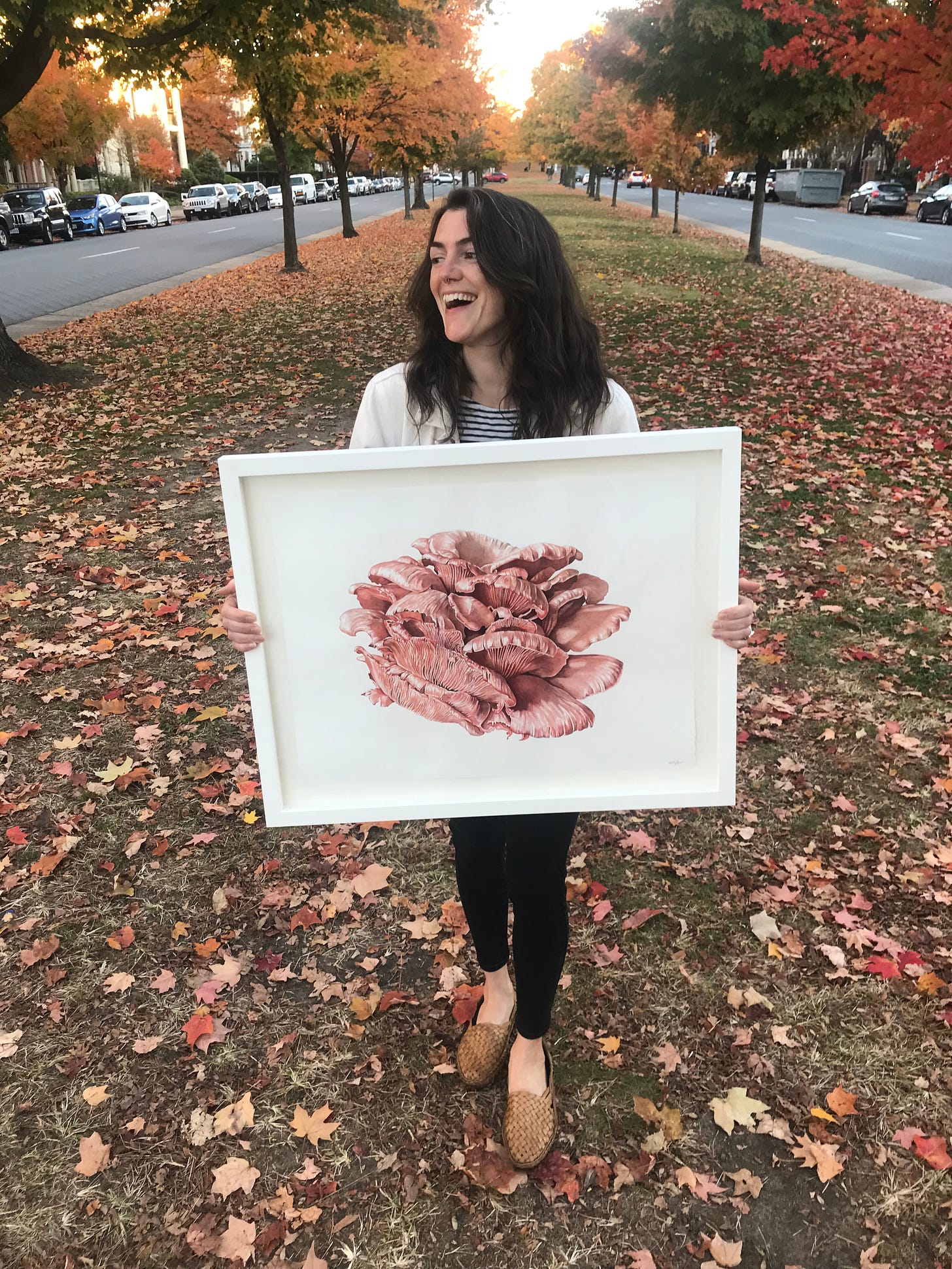 Me standing and smiling holding the finished framed painting, with autumn leaves and trees behind me in the background. 