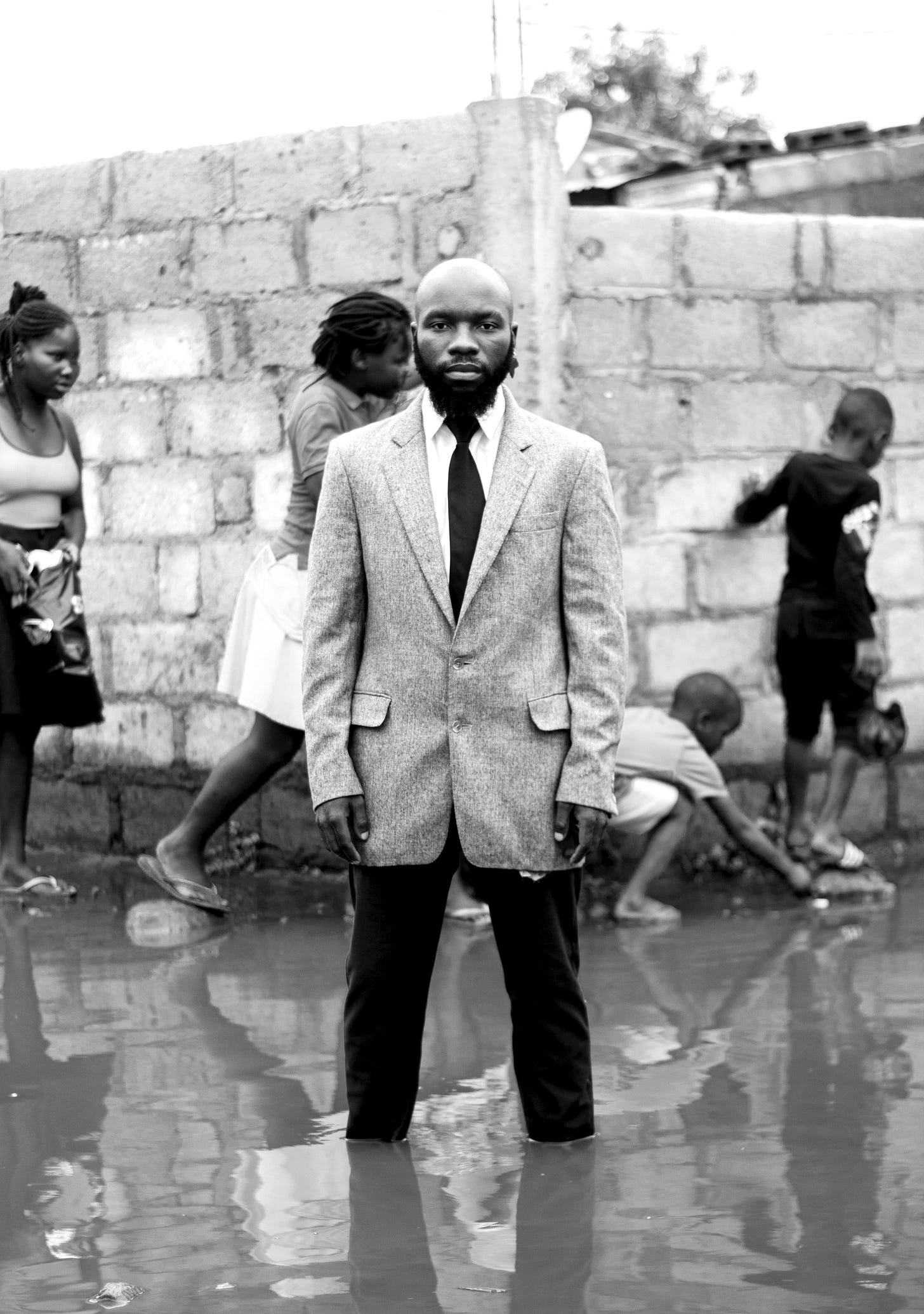A man dressed in a suit standing in the middle of a puddle, people in the background are stepping on stones trying to cross the puddle