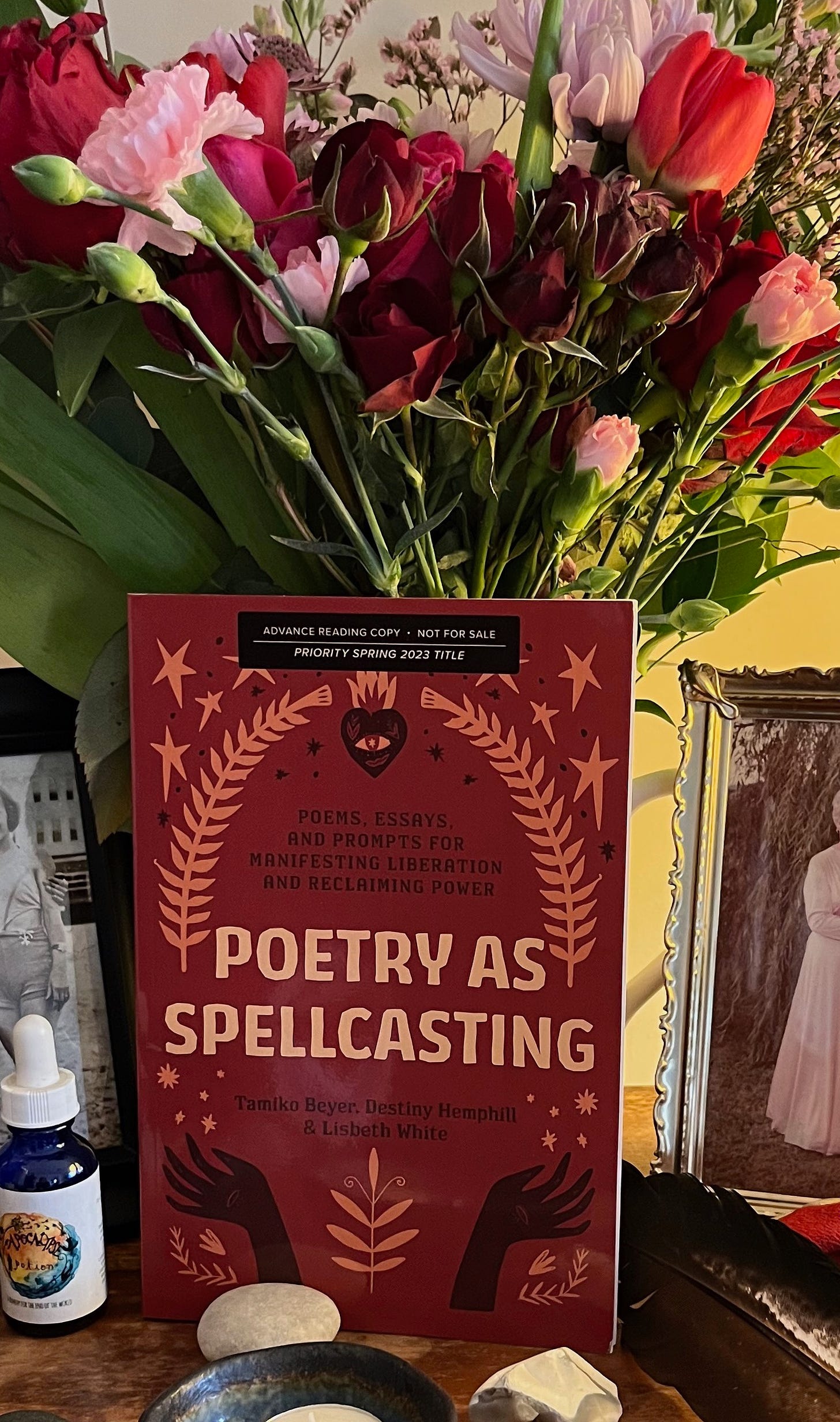 An advanced reading copy of Poetry As Spellcasting is placed on an altar  in front of a bouquet of roses, tulips, chrysanthemums, and carnations. The reds and oranges of the books match the colors of the flowers.
