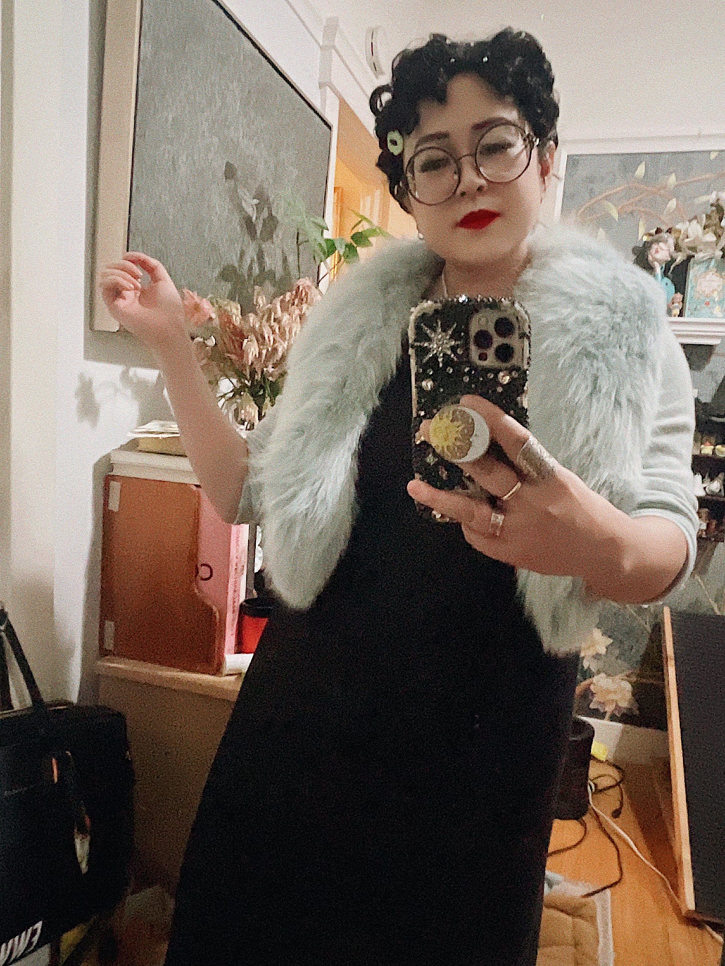 A person wearing glasses, red lipstick, and a light blue faux fur shrug over a black dress takes a mirror selfie. Their hair is styled in tight curls with decorative pins.