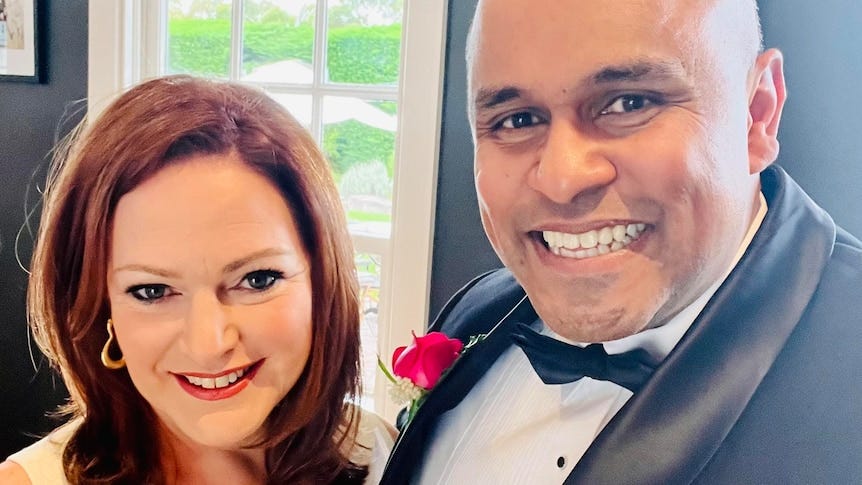 Emma wears a white sleeveless dress and stands closely to Raj who is in a tuxedo with a red rose on his lapel
