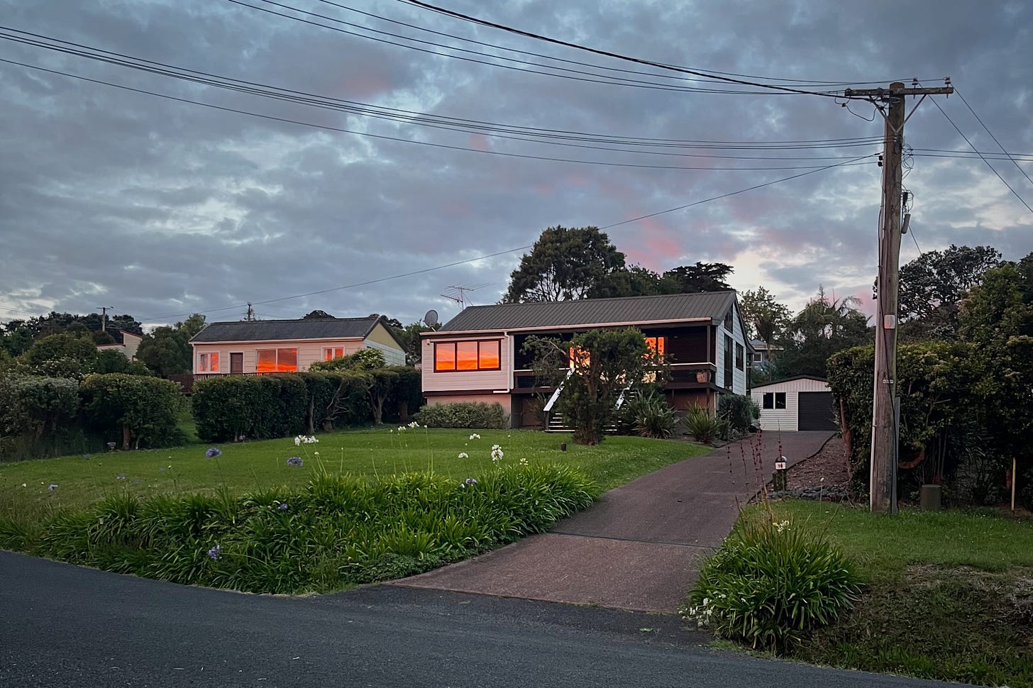 the sun is setting and the red light is reflected in the windows of a 1980s New Zealand house set off a road on a sloping lawn, agapanthus plants at the verge
