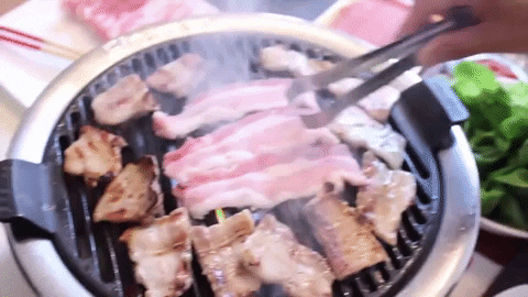 A hand holding tongs turns meat strips on a grill.