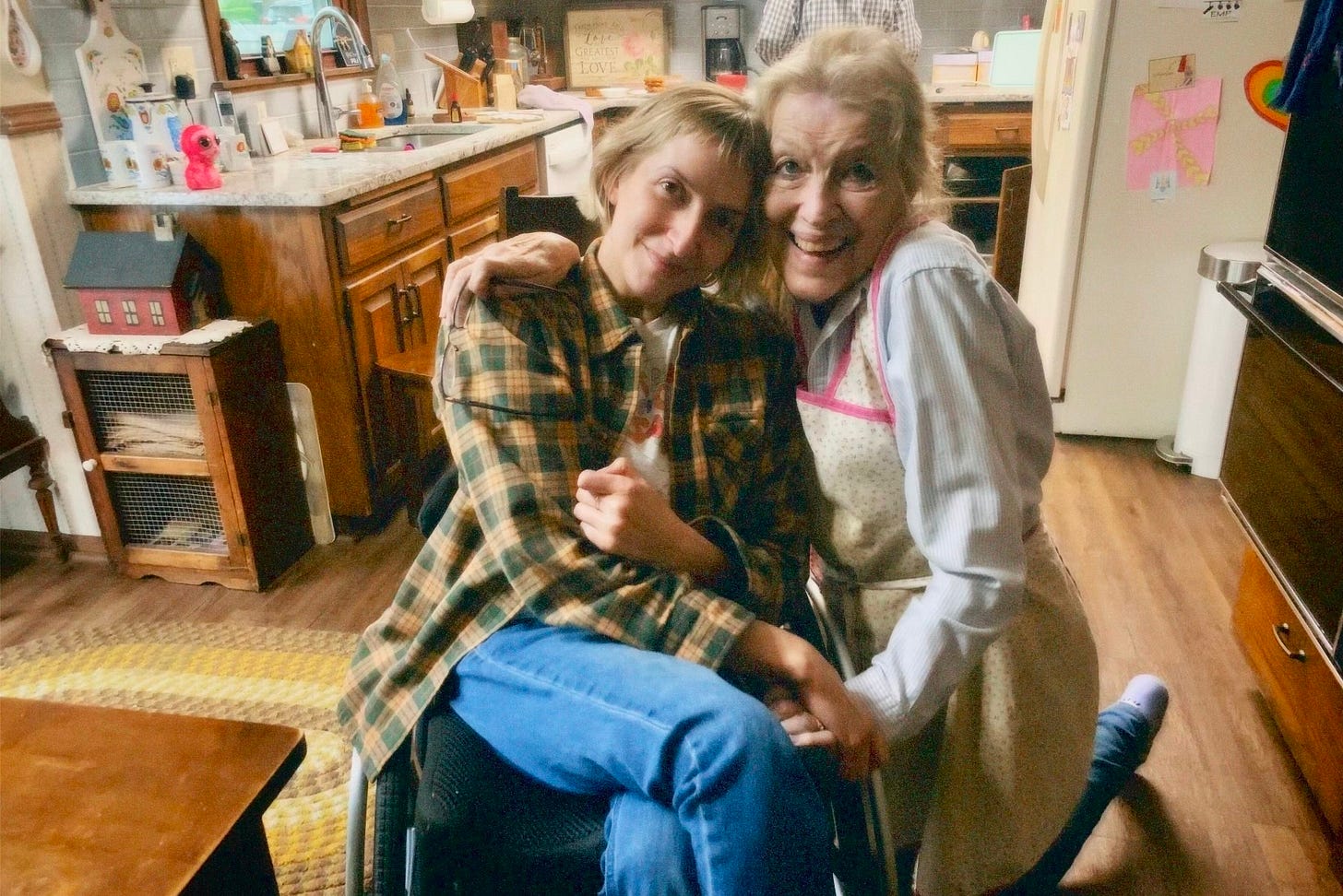 A 39-year-old Rebekah sits in her wheelchair next to her 75-year-old mother who is on her knees so they are level. Rebekah tilts her head toward her mom's, her temple against her mom's cheek. They are holding hands, making a heart shape with their arms. Behind them, the kitchen in the house Rebekah grew up in. A 39-year-old Rebekah sits in her wheelchair next to her 75-year-old mother who is on her knees so they are level. Rebekah tilts her head toward her mom's, her temple against her mom's cheek. They are holding hands, making a heart shape with their arms. Behind them, the kitchen in the house Rebekah grew up in.