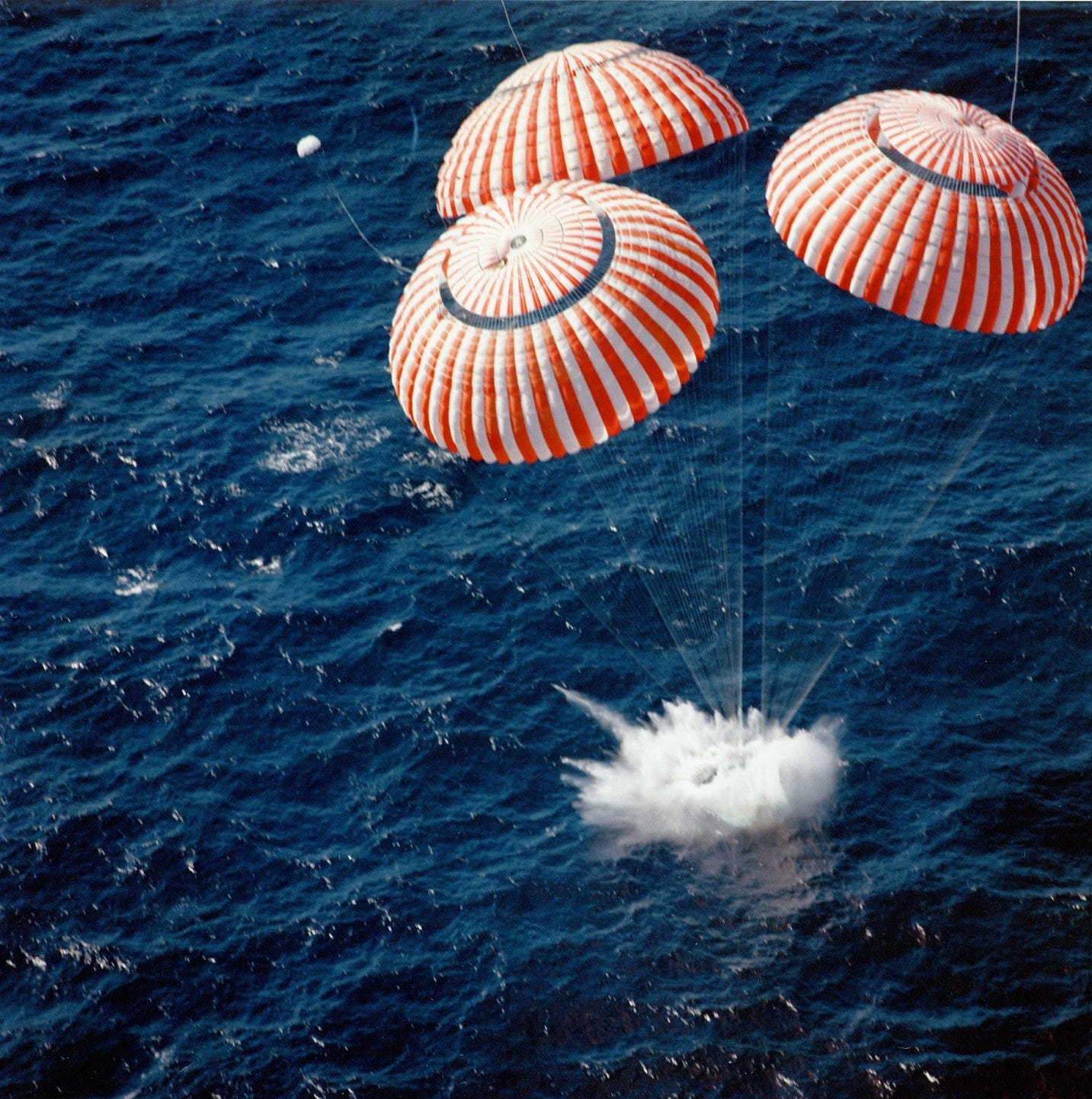 Three red and white parachutes float above a white splash in a blue ocean.