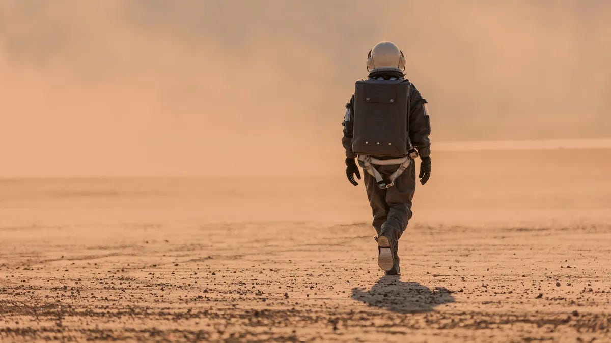 A person in an astronaut suit walks across a reddish desert landscape. 