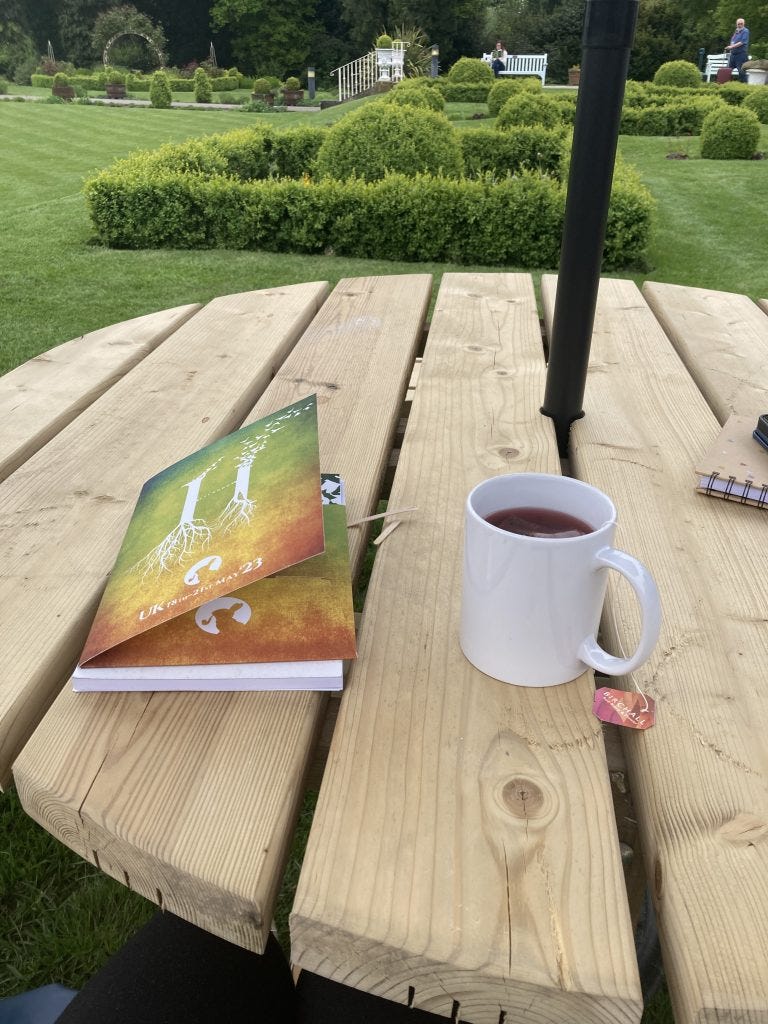 A round wooden picnic table in the gardens on the conference centre. On the table is the ombre (orange to green) conference folder for Hutchmoot 2023 and a white mug. In the white mug is a purple herbal tea.