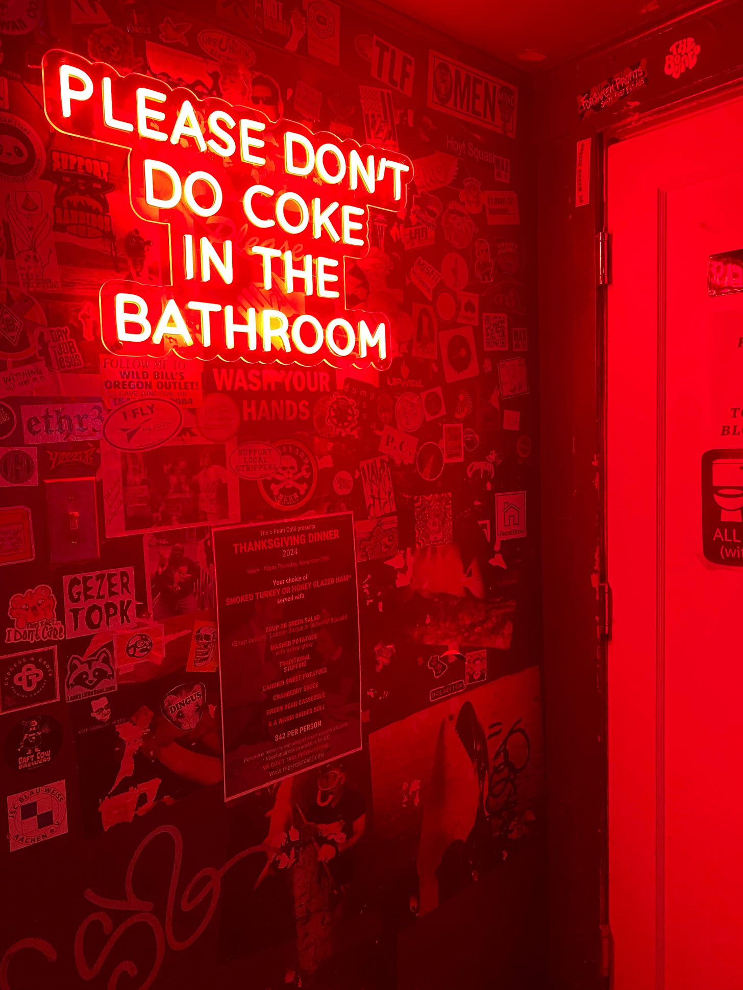 Wall outside the bathroom at The 5 Point, with many posters and stickers on the wall, which is lit by a neon red sign that says 'PLEASE DON'T DO COKE IN THE BATHROOM'