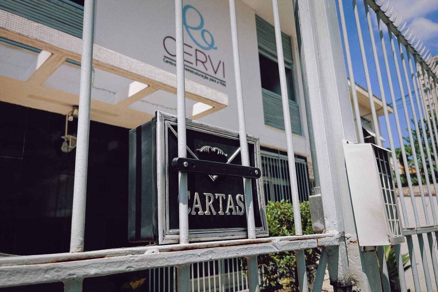 A white building with a sign that says CERVI, seen through a tall gate. Bright blue sky is visible in the background.