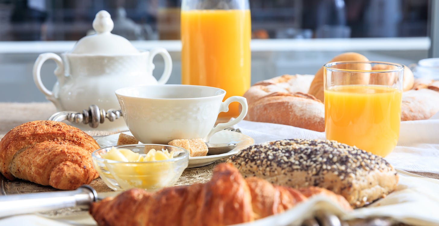 Breakfast table. There’s bread, croissants, butter and glasses of orange juice, as well as a little white teapot and a white teacup.