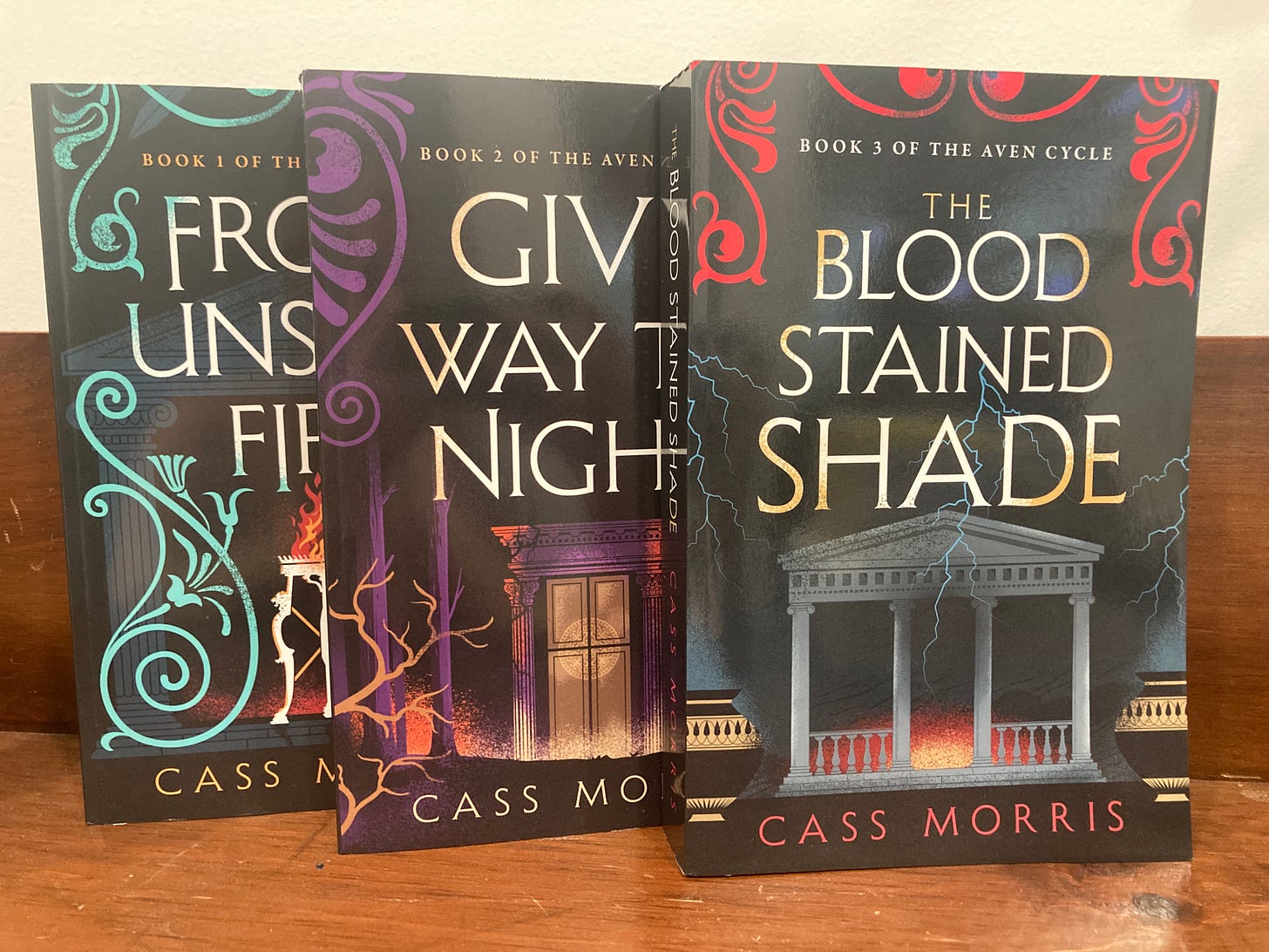 Paperback editions of FROM UNSEEN FIRE, GIVE WAY TO NIGHT, and THE BLOODSTAINED SHADE, standing upright and overlapping on a wooden table