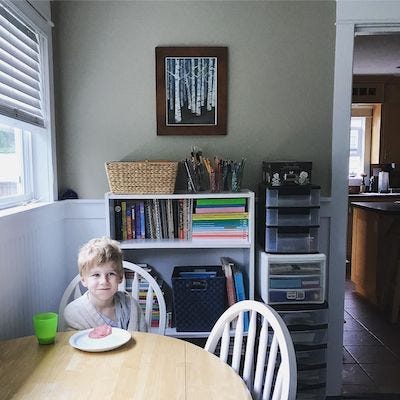 A small boy sits at a round table, smiling, wrapped in a blanket. His breakfast and a green cup of milk are in front of him. Behind him: the bookshelves and supply drawers of my studio.