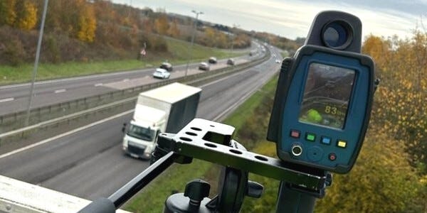 Tripod-mounted speed camera positioned on a bridge overlooking traffic on a fast road below