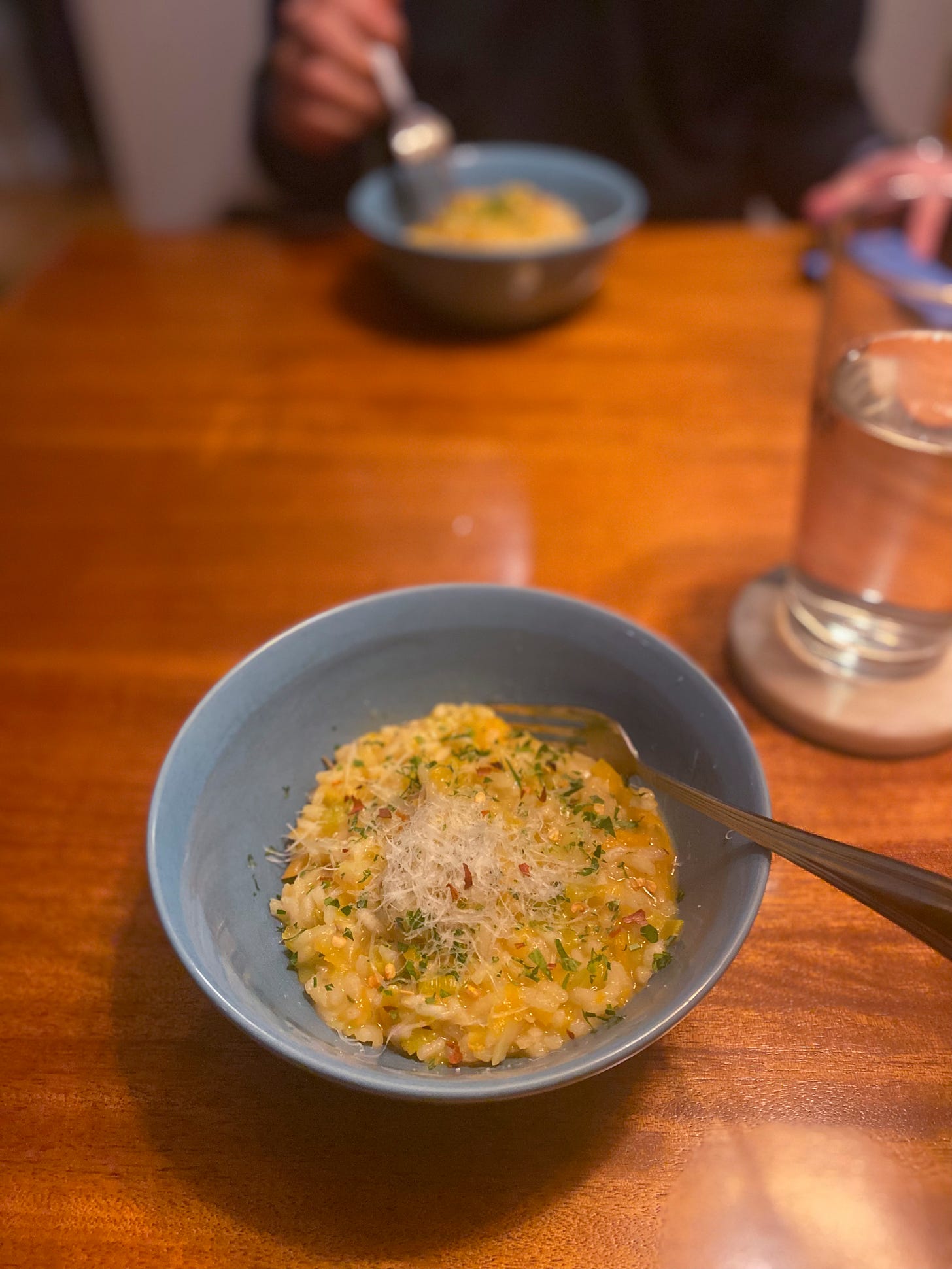 A blue bowl of the risotto described above, coloured lightly orange from the squash and dusted with parsley, chili flakes, and a little pile of grated parmesan. Jeff's fork is poised over his bowl in the background.