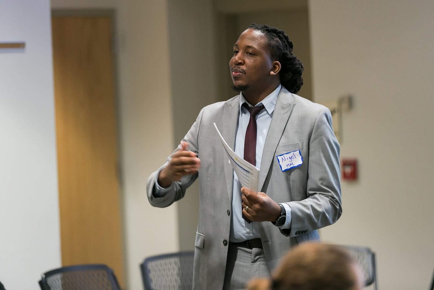 Nigel standing in a conference room, talking to a group that is not pictured, wearing a grey suit, with his braids tied back behind his head.