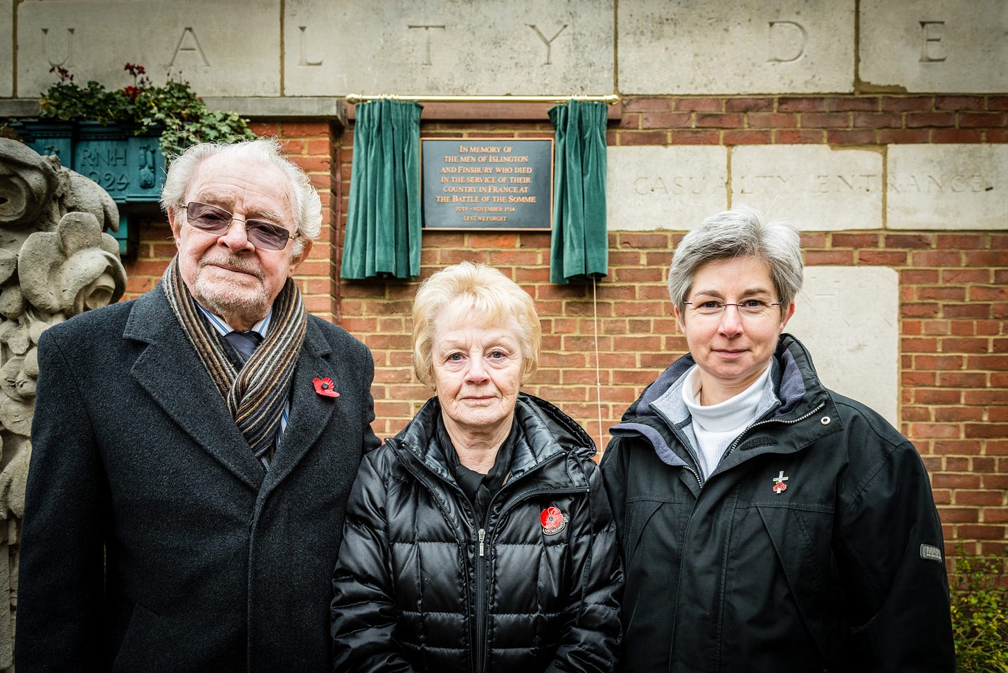 A man and two women stand in front of a brick wall with a commemorative plaque. © Em Fitzgerald Photography 2016. All rights reserved.