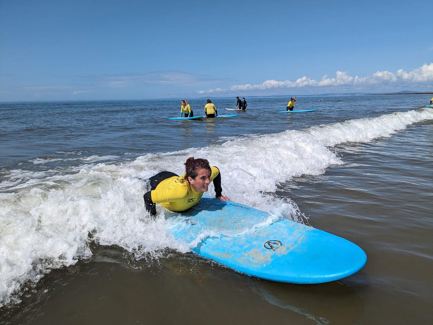 A woman in a black wetsuit and yellow safety top is lying on a surfboard, enjoying the wave beneath her. Her face is a picture of delighted concentration as she flows with the water