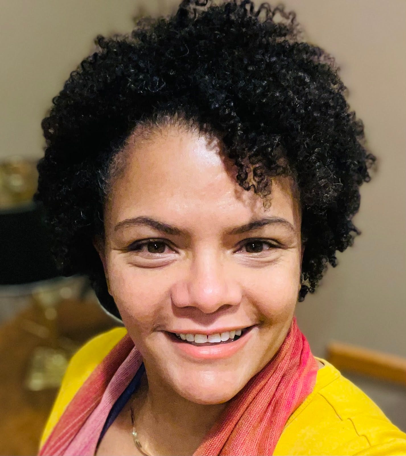 A photo of a smiling Black woman with mid-length curly hair. She is wearing a yellow shirt and orange scarf.