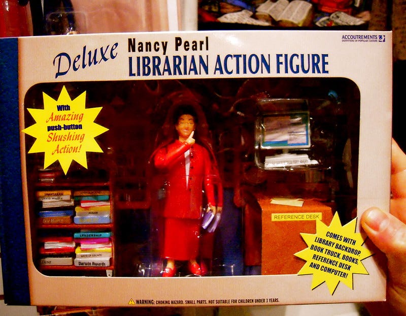 Photograph of a person holding the Deluxe Nancy Pearl Librarian Action Figure complete with the book cart, computer station and reference desk backdrop in its original packaging.