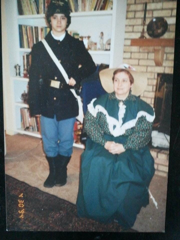 The author as a young, very androgynous-appearing person dressed as a Union Civil War soldier, posing next to their mother, who is seated and dressed in a green patterned dress and bonnet that she made to be period accurate.