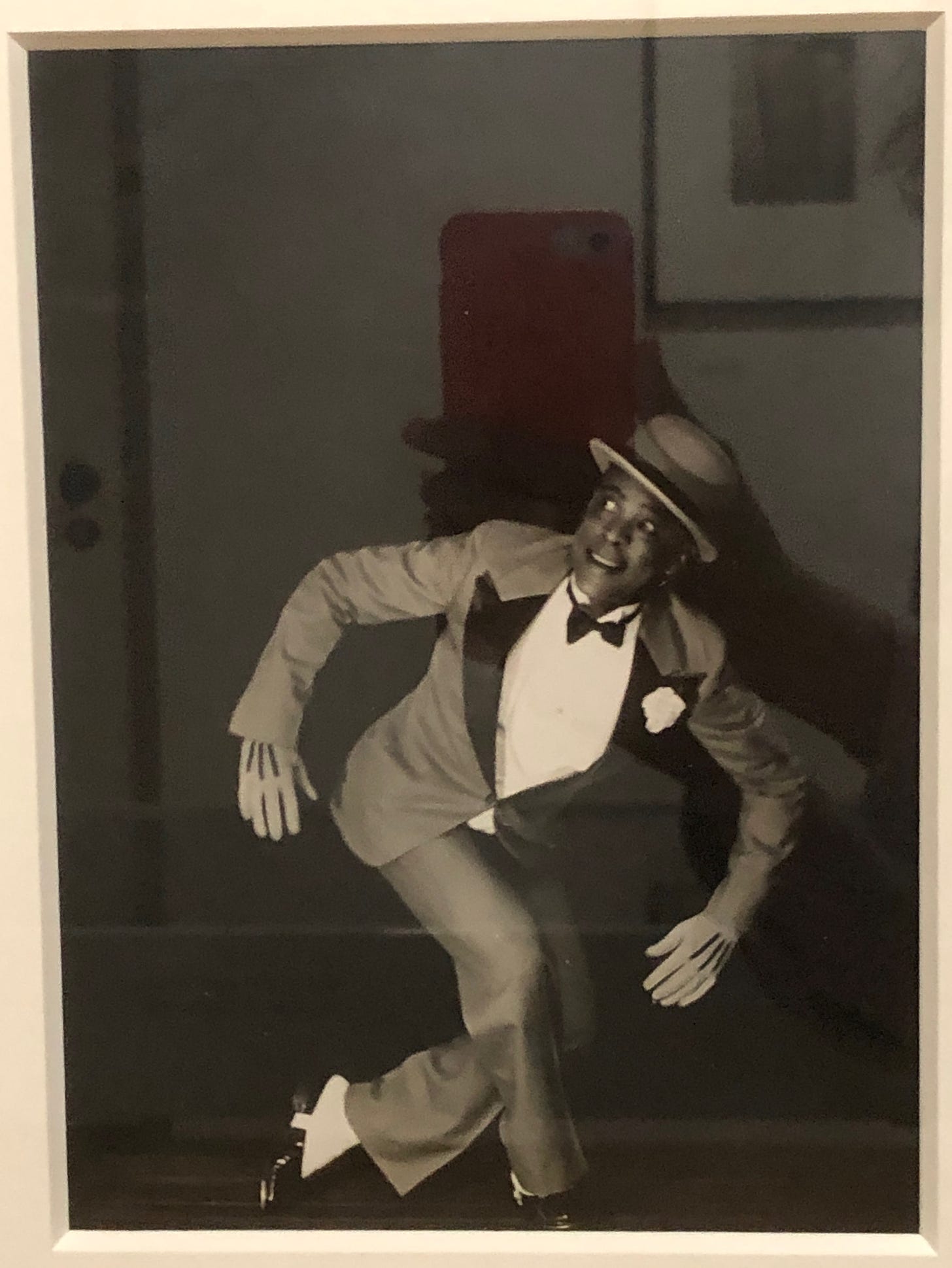 A man in a suit, bowtie and bowler-ish hat in tap-dance action: left knee fully bent, right leg stretched out straight in front, left arm outstretched and pointing downwards, and right hand in the air, raising his hat.