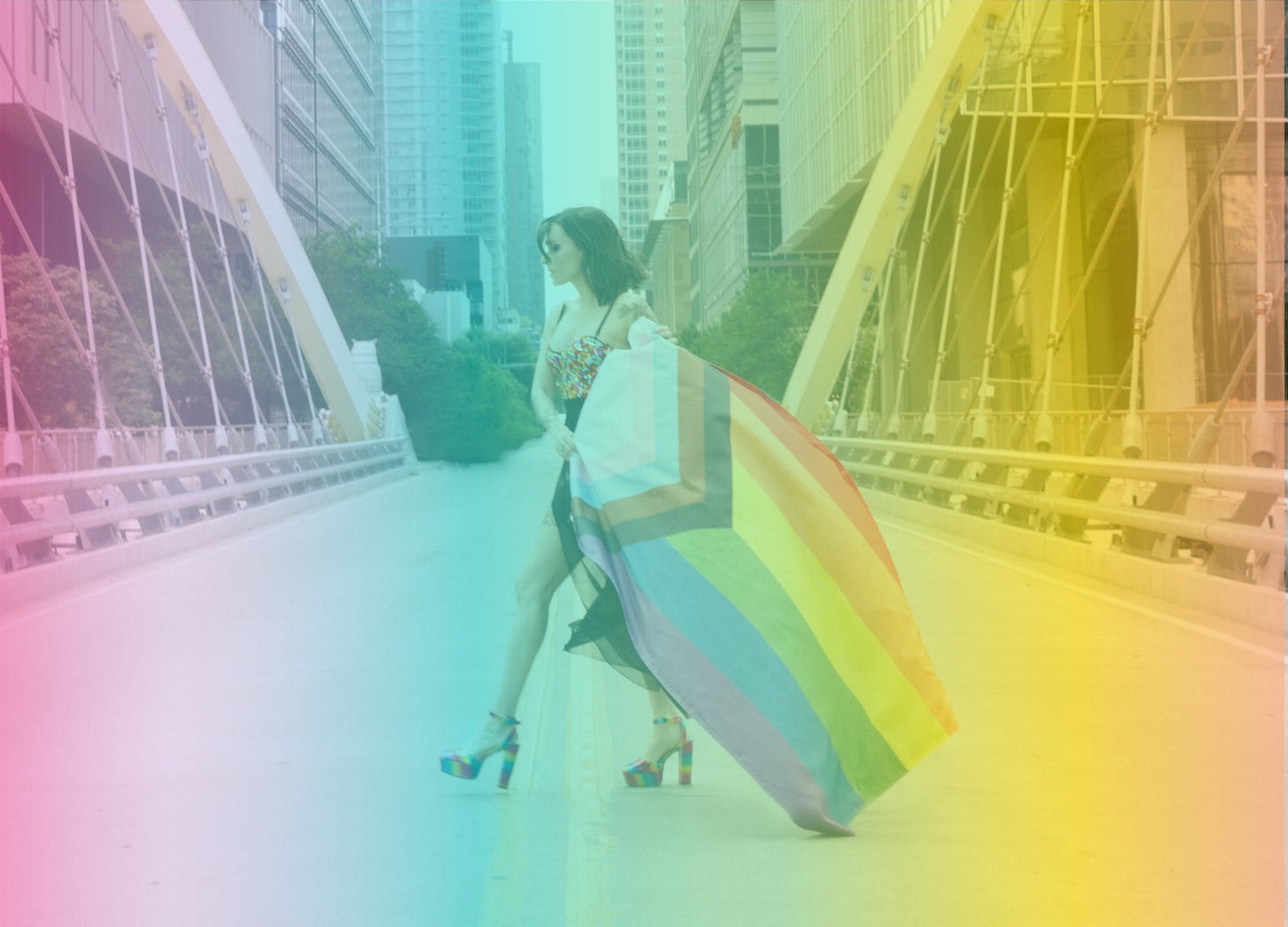 A rainbow-filtered photo of Shohreh crossing the road on a bridge while holding out a Progress Pride flag and wearing rainbow platform heels