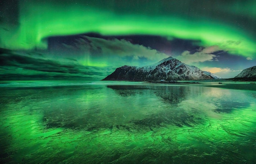A wide view of a bright-green aurora above a snow-covered mountain, appearing like a shimmering curtain in the sky.