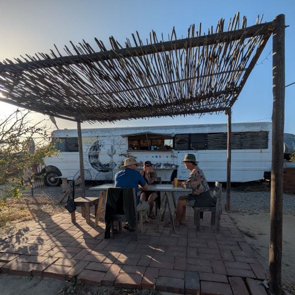 three people sit at a rustic outdoor table on a paving stone patio with a thatched roof overhead. Behind them is a long white bus with a serving window in the side.