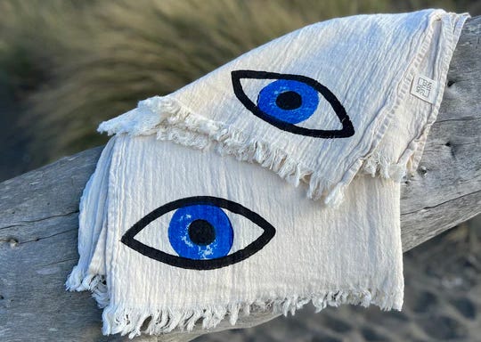 Two evil-eye design peshtemals are draped on a driftwood log in front of a sand-and-sea-grass scene.
