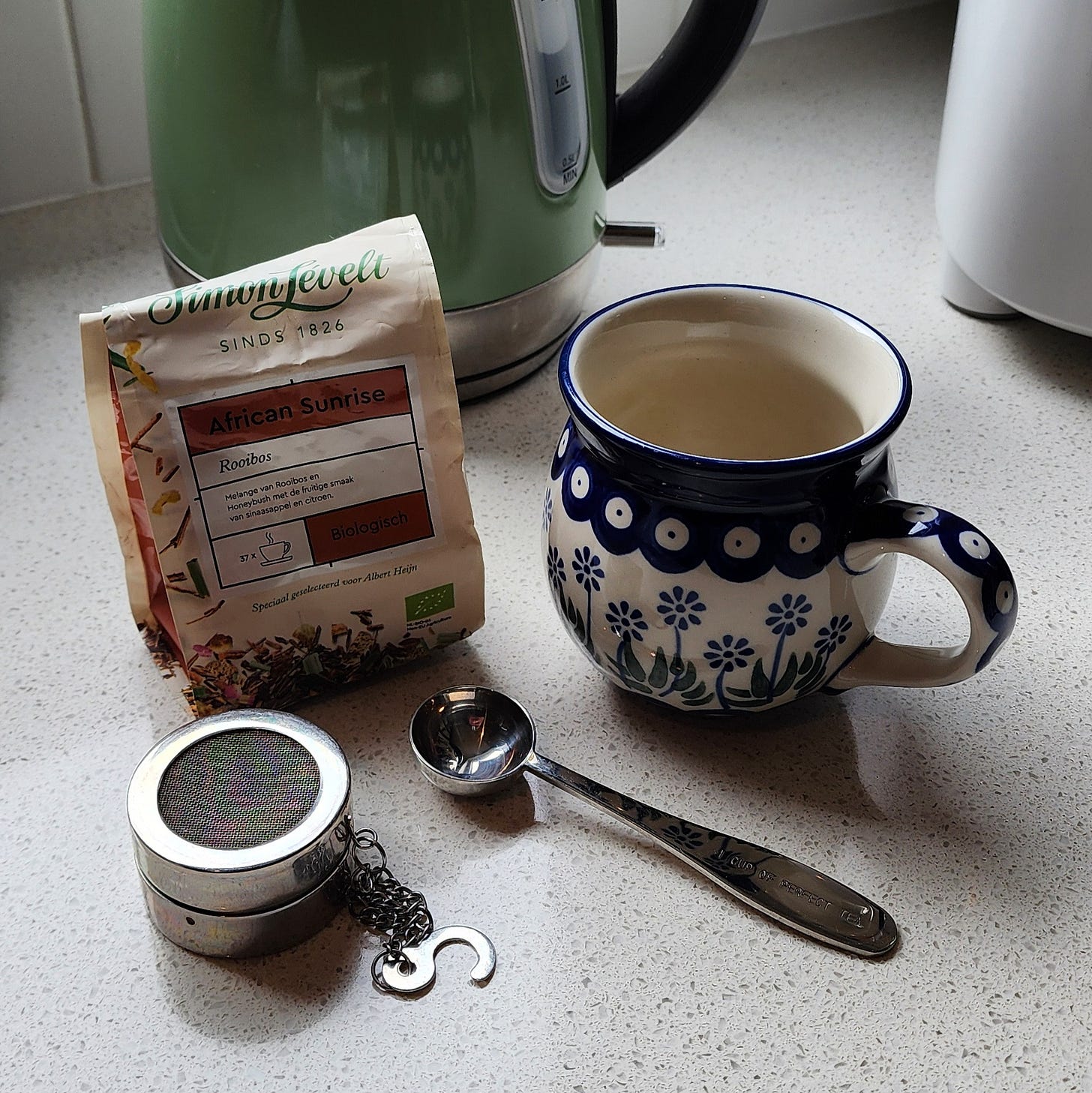 Ingredients and tools for making a cup of tea sit on a kitchen bench. An infuser and a measuring spoon are in the foreground, with a Polish pottery mug, and a packet of Simon Levelt 'African Sunrise' tea. The bottom of a green electric jug is in the background.