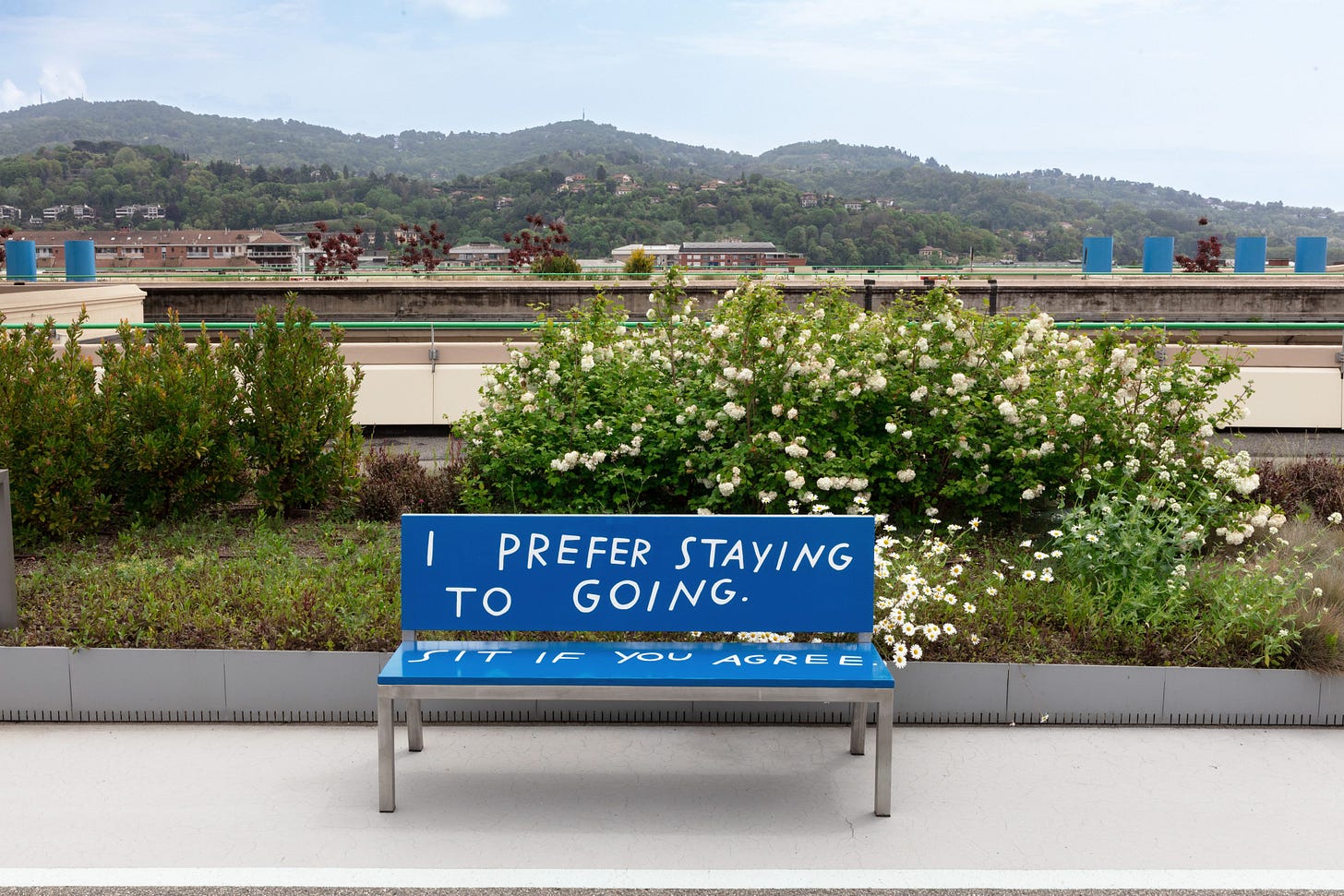 A photo of a park shows a blue bench with silver legs. White text on the bench’s back rest says I PREFER STAYING TO GOING. On the seat of the bench, it says SIT IF YOU AGREE.