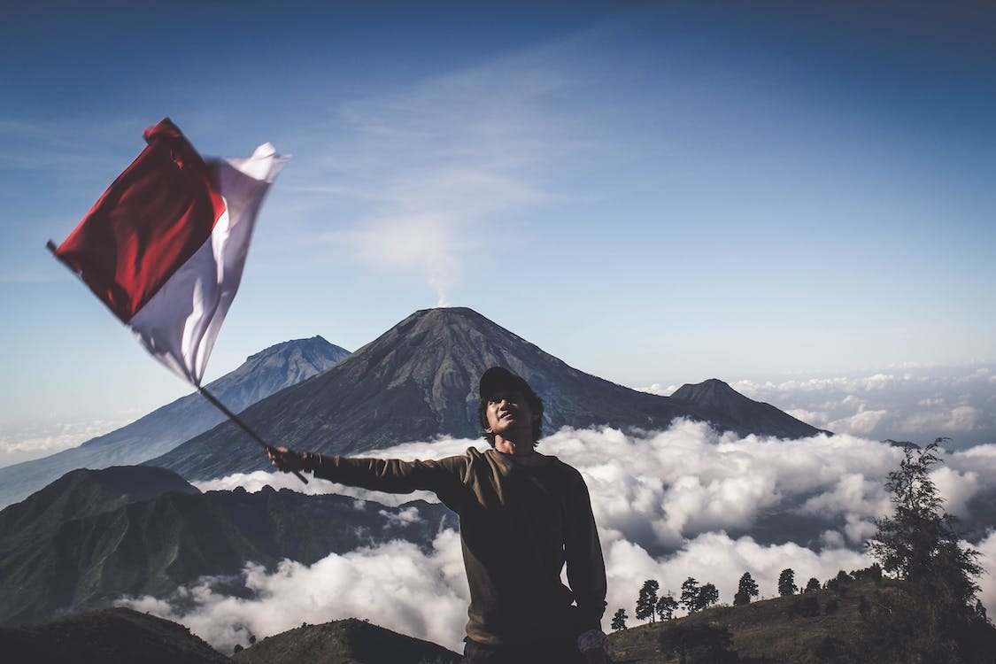 Free Man Wearing Black Crew-neck Sweater Holding White and Red Flag Standing Near Mountain Under Blue and White Sky Stock Photo