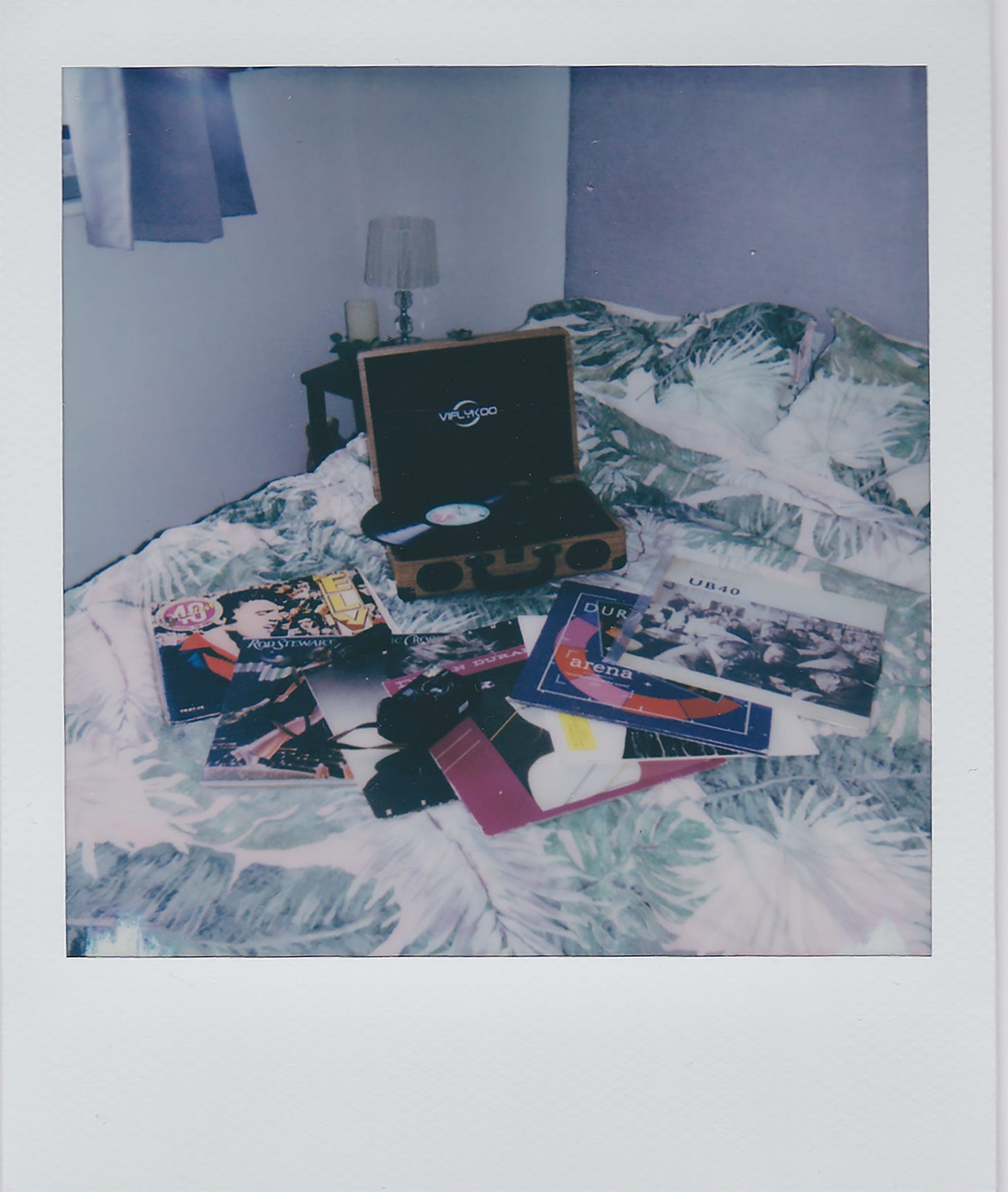 Polaroid picture of record player sitting open on a bad with a record on the turntable, various records fanned out on the bed in front of it.