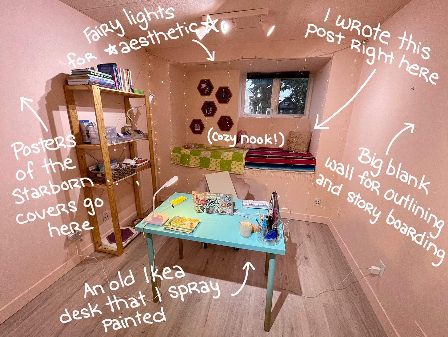 Image shows a room with pink walls. Wooden shelves sit against a wall, a teal desk sits in the middle of the floor, and a blanket-covered nook sits under a window at the back of the room. There is white writing on the image pointing out various features of the room.