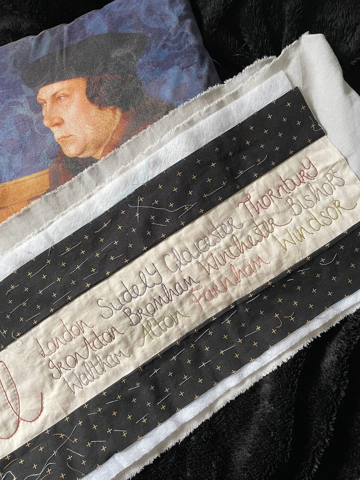 A quilted piece in black and gold, with a cream panel containing place names quilted on to it. A cushion featuring Thomas Cromwell is behind it.