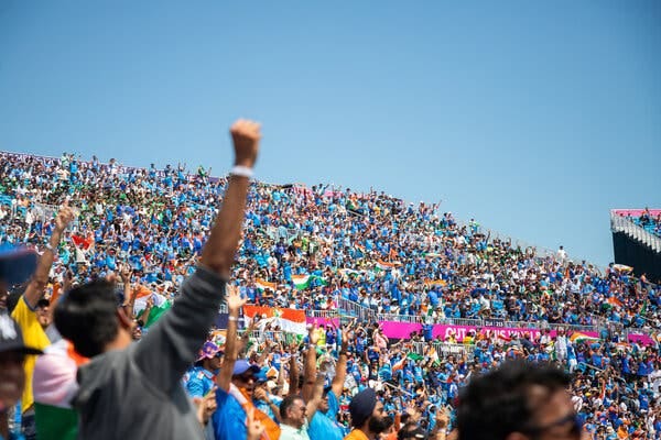 Fans raise their arms and cheer in a crowded stadium under a clear blue sky.