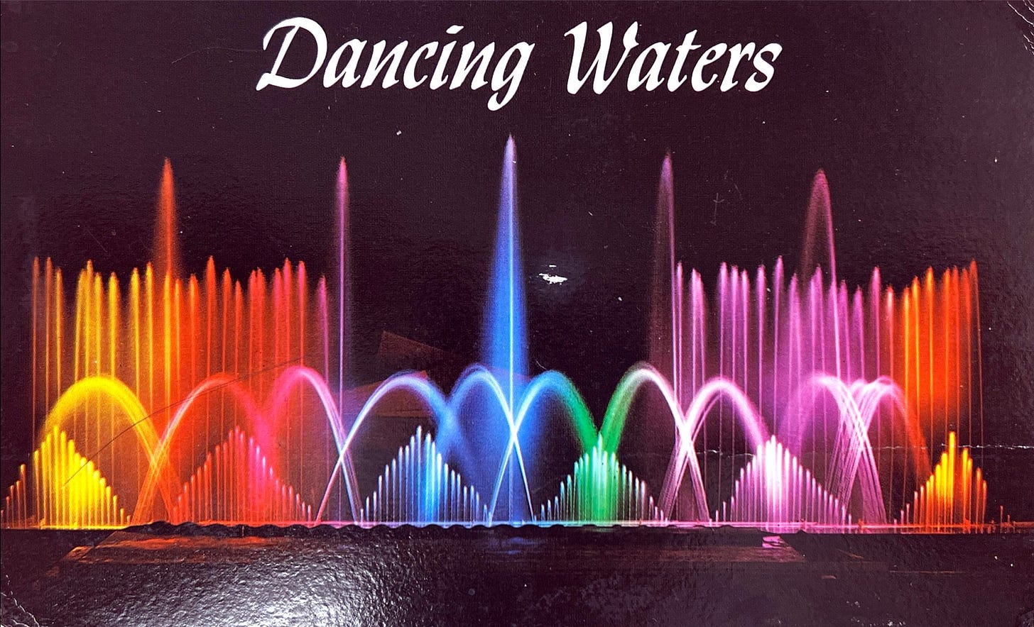 Photo of water fountain lit up in rainbow colors with the title "Dancing Waters."