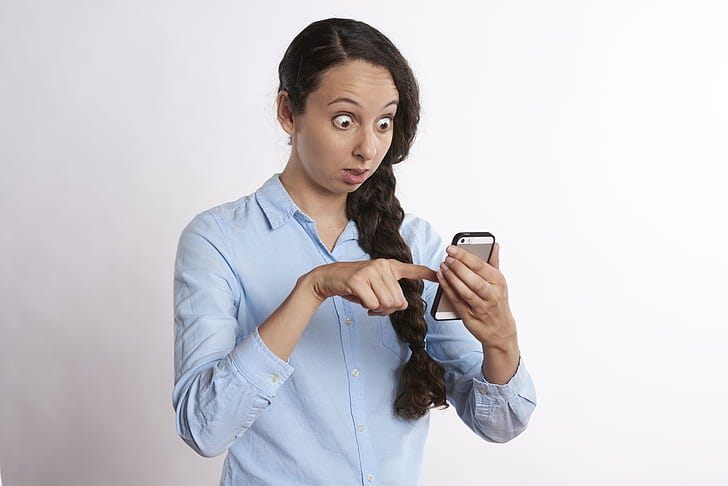 A woman wearing a blue button-up collared long-sleeved shirt holding a gold iPhone 5s with black bumper case, staring at the phone in shock