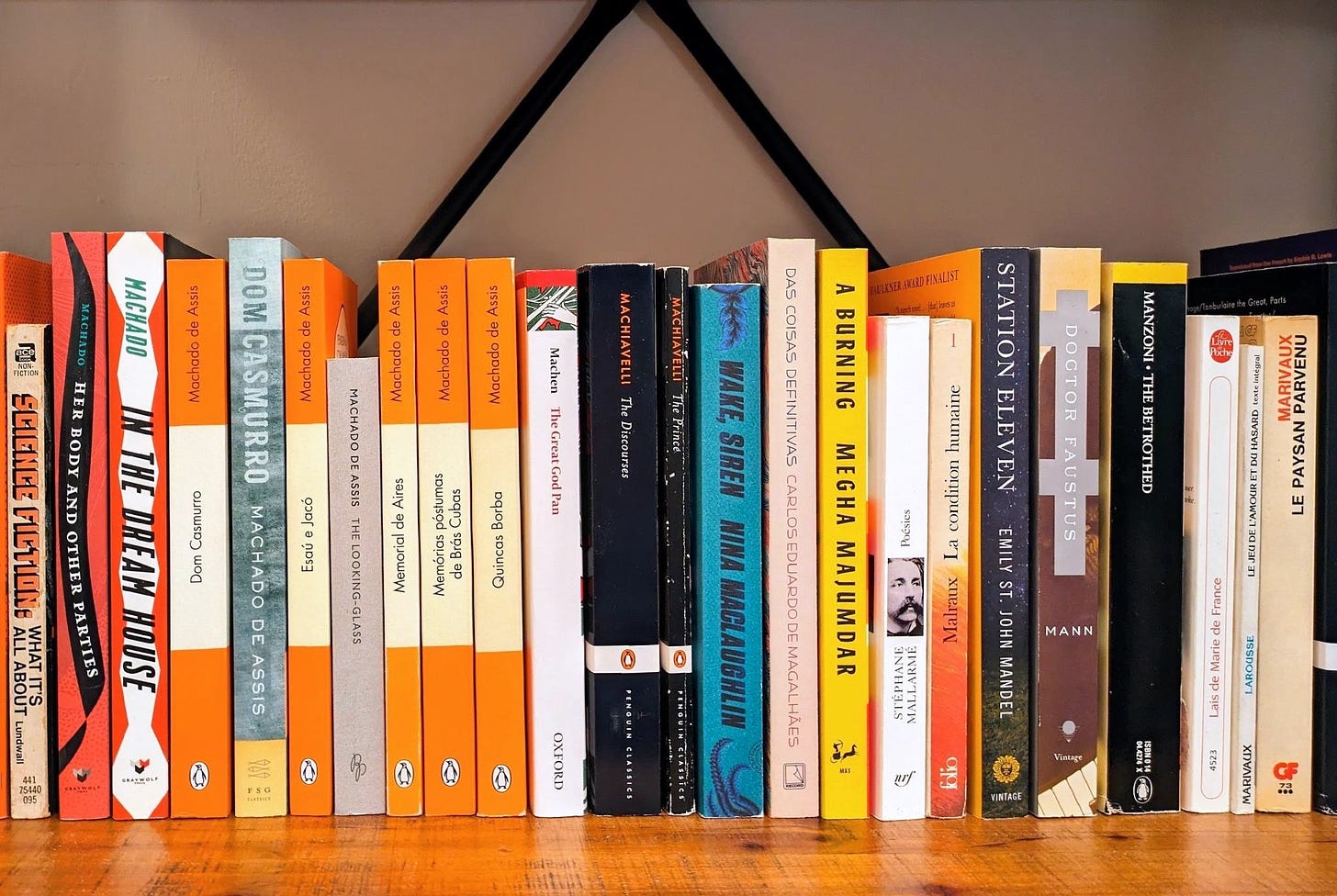 Photo of a section of a bookshelf with various bookspines visible, some in Portuguese, some in French, the majority in English. There's a concentration of bright orange spines at left, all by Machado de Assis.