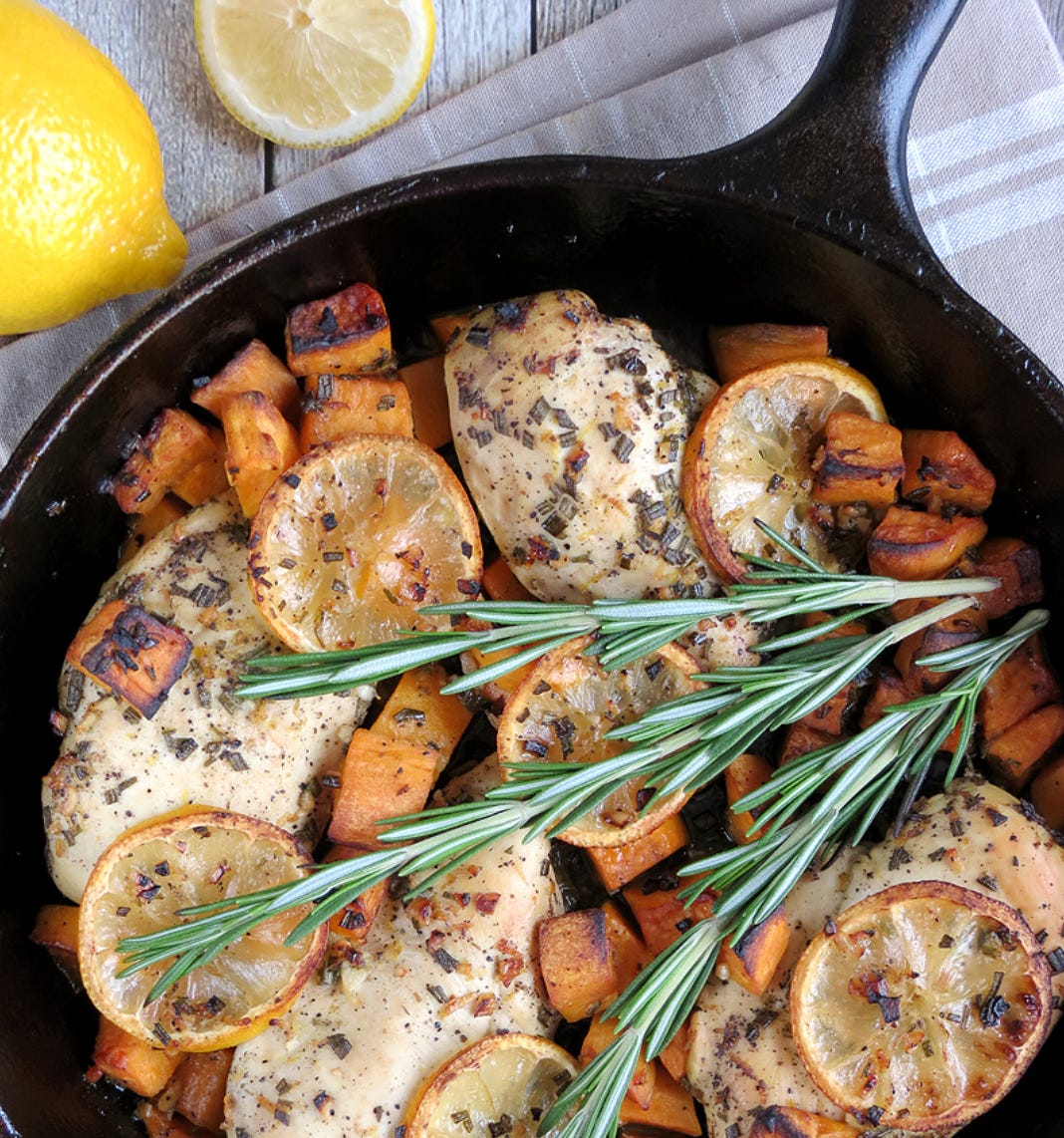 Chicken and sweet potatoes in cast iron skillet, with lemon slices and rosemary sprigs.