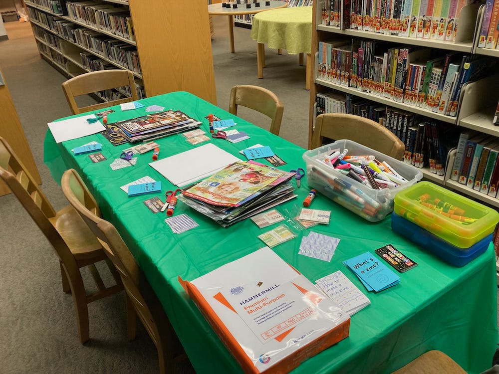 A table with zine-making supplies: paper, glue sticks, scissors, magazines, and scrapbook paper. The table is inside a library.