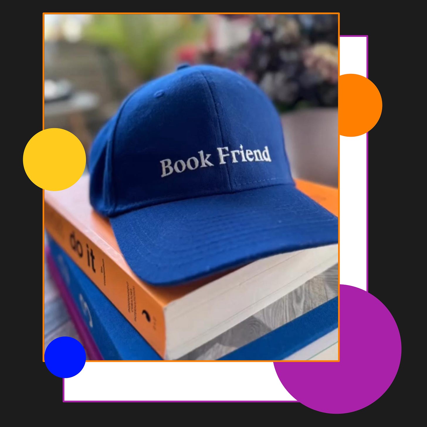 A blue baseball cap reading “Book Friend” on top of a pile of books.