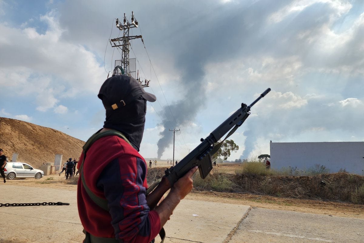 A person holding a rifle on a road, with plumes of smoke in the distance.