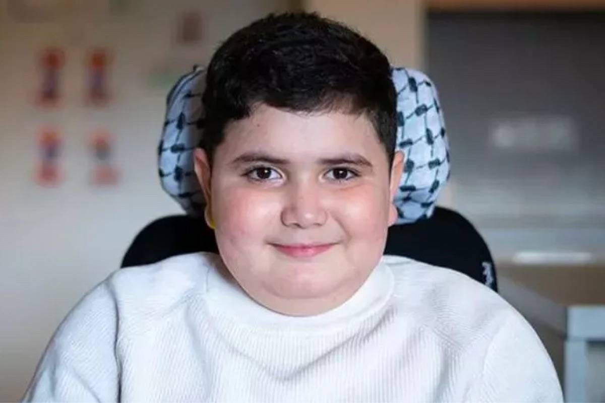 A wheelchair using boy is smiling at the camera. The headrest of his powerchair is wrapped in a keffiyeh.