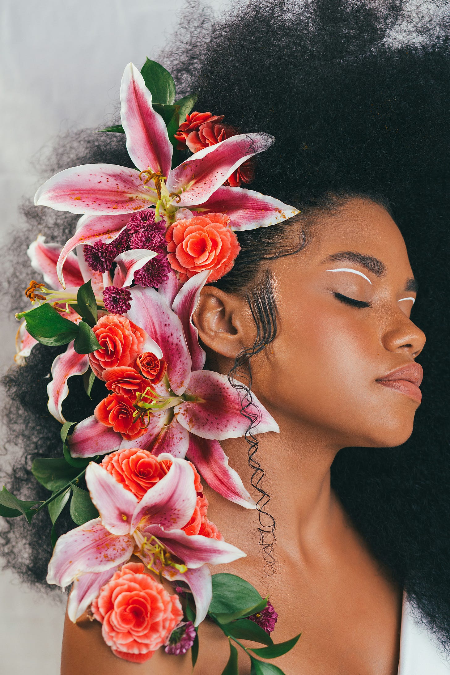 A light gray background behind a Black woman with warm brown skin. She has dark natural hair that is full and to one side. She has colorful tropical flowers trailing down over her shoulder on the other side. Her eyes are closed and she has a white line on her eyelid between her eyebrow and lashline.