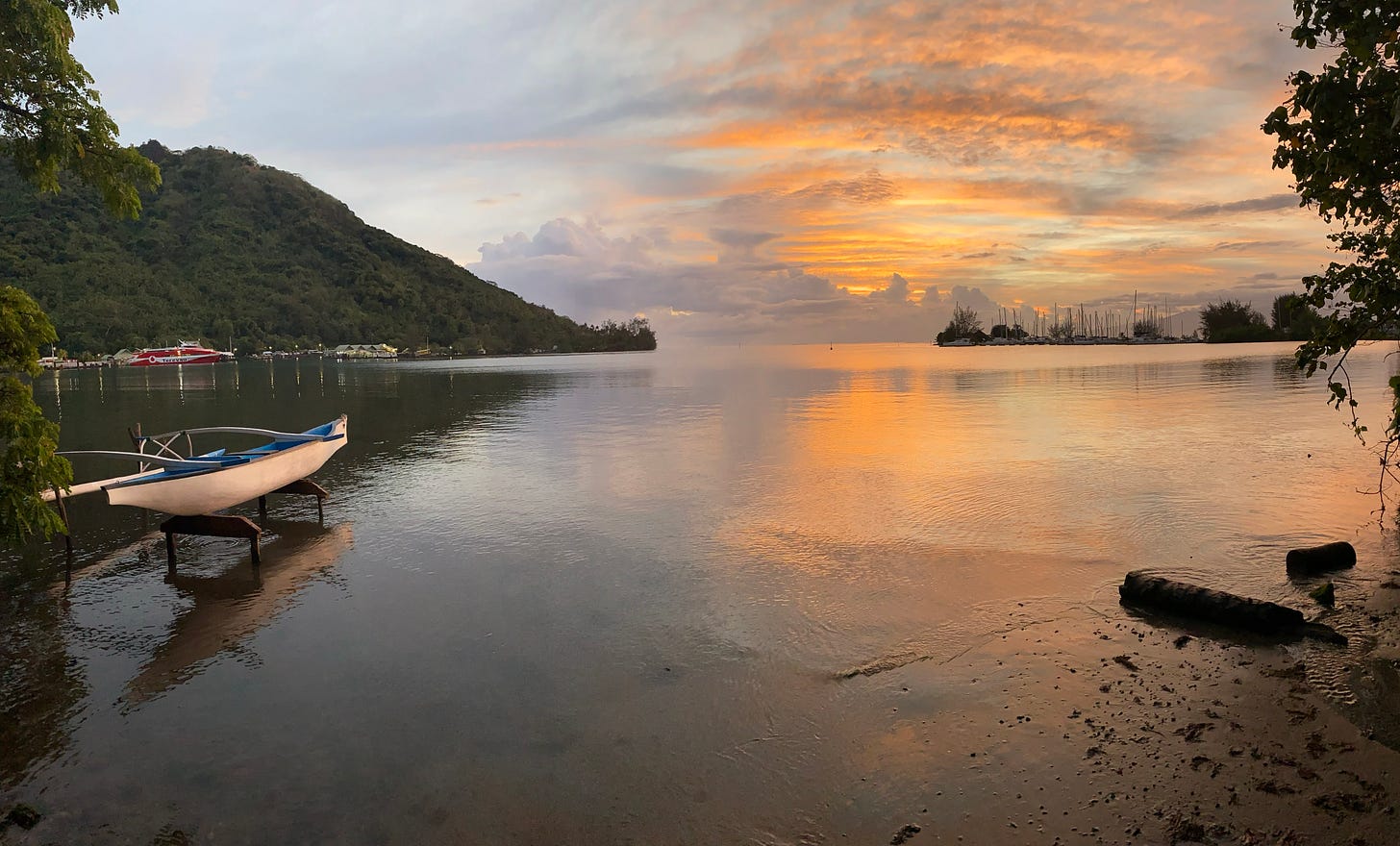 A sunrise over a bay with a mountain and marina in the background. In the foreground is a blue and white pirogue. 