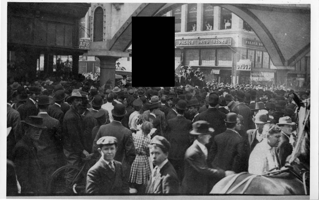 A redacted photo showing the lynching of Allen Brooks, a Black man, which took place in Dallas, Texas on March 3 1910. The crowded street scene shows thousands of people, including what appears to be two young children look towards the camera in the foreground.