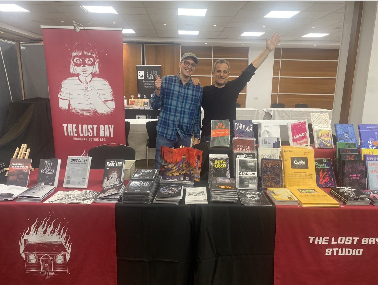 Chris Airiau and Iko behind The Lost Bay stall at Dragonmeet, lots of zines