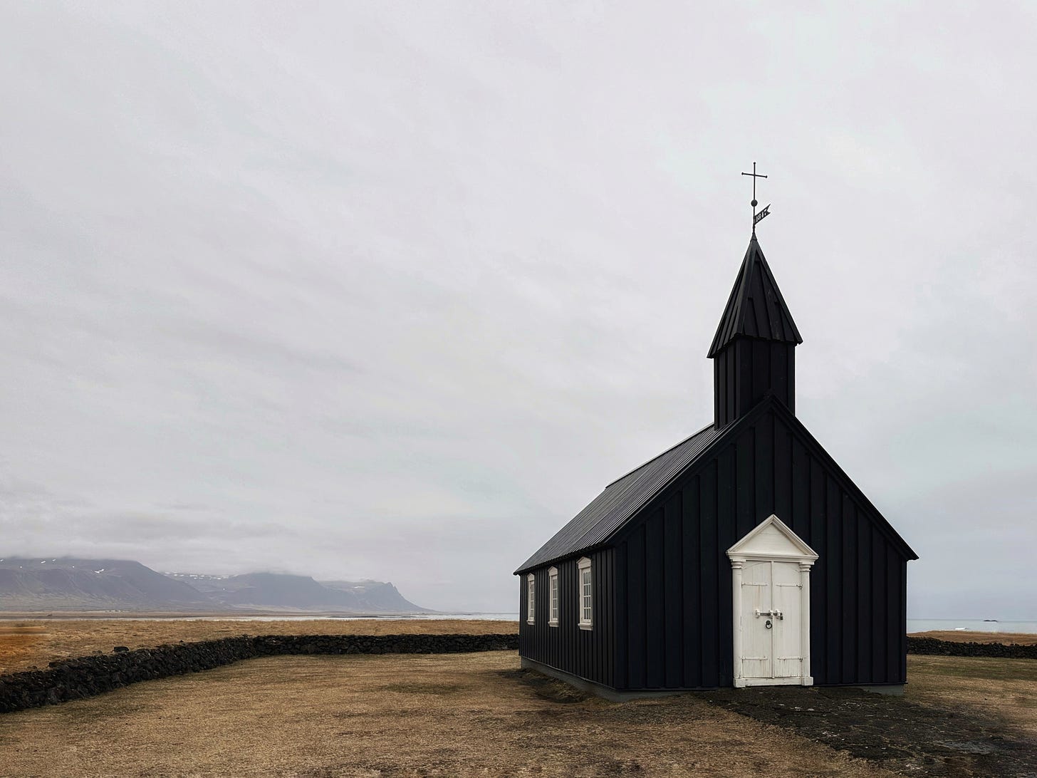 Búðakirkja - The Black Church of Budir a small Lutheran church painted black with white trimmed windows and door.  This church is alone on a moss covered lava field with mountains in the distance and the sea on the right.  The ground is golden brown and the sky is filled with light gray clouds.