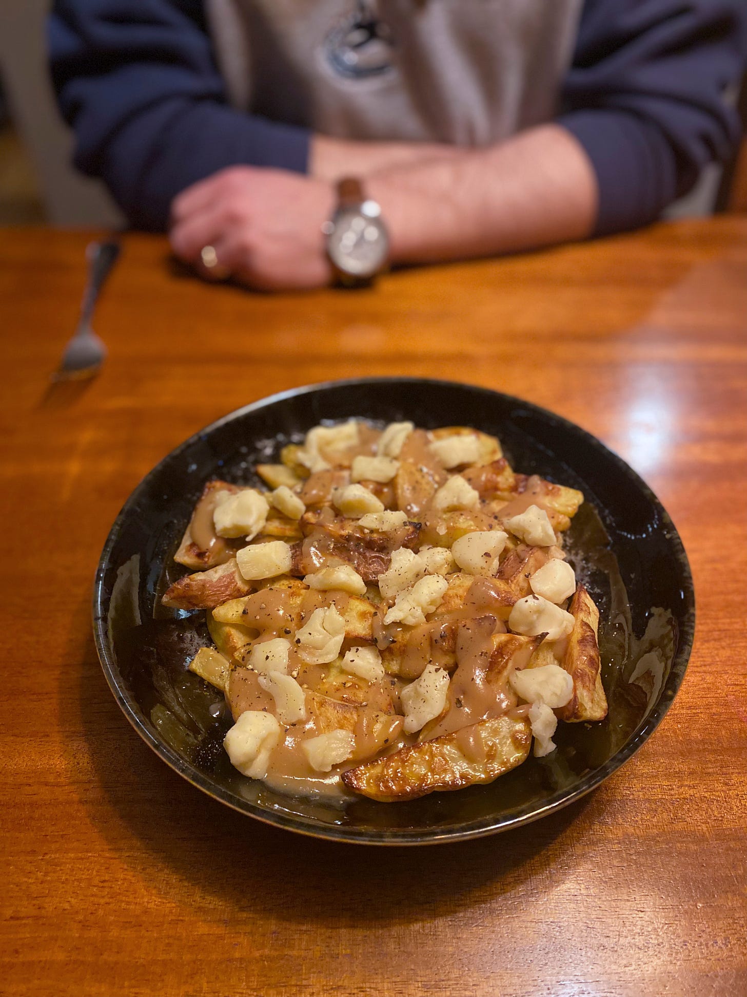 A black serving dish of poutine: small wedge fries covered in gravy, cheese curds, and black pepper. In the background, Jeff sits at the table, his arms resting on it next to his fork.